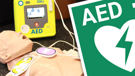 Gelber Defibrillator, Pads auf einer Demonstrationspuppe. Im Anschnitt das grüne Hinweiszeichen für AED-Geräte.